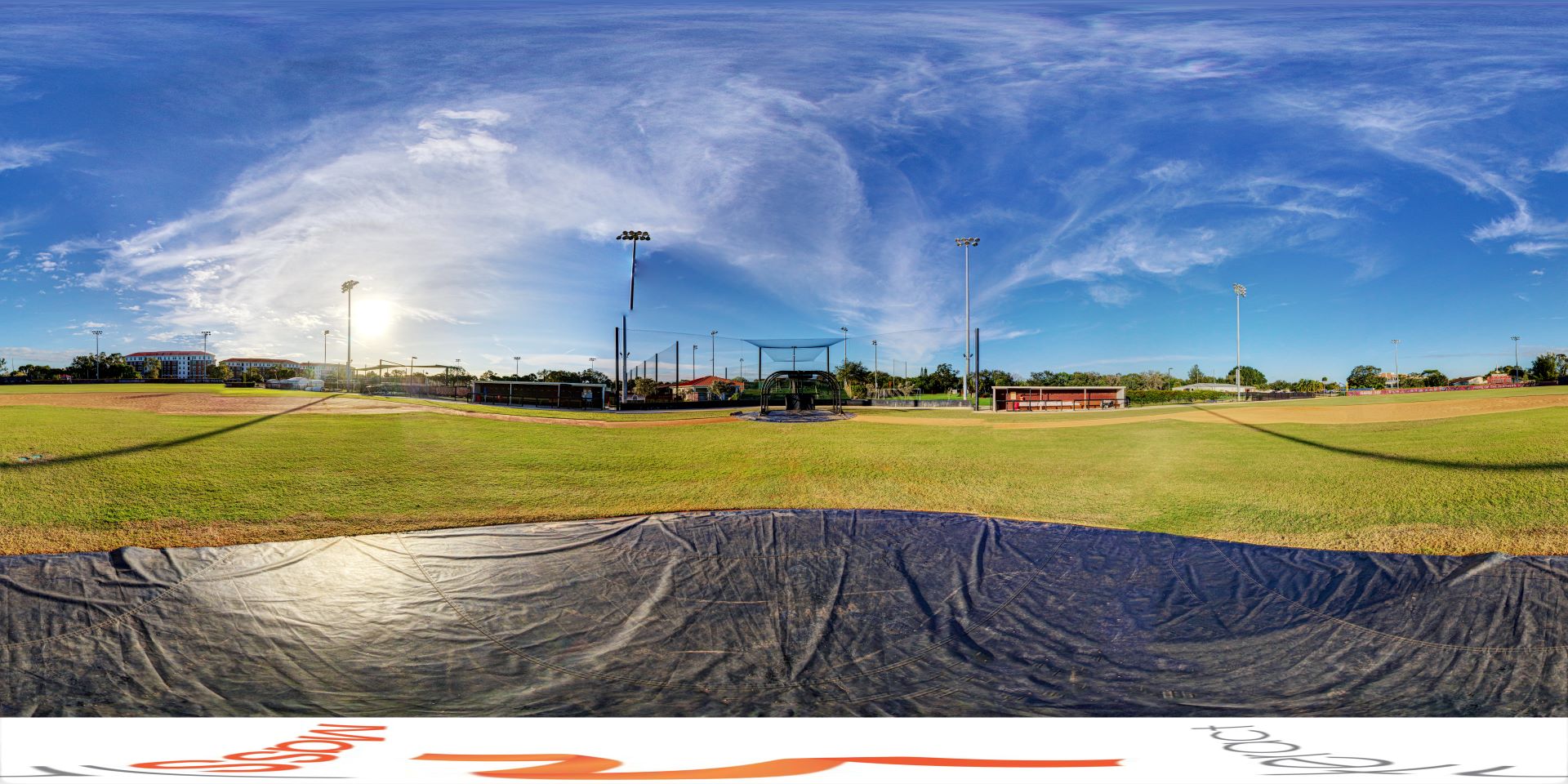 A panoramic view of Andy Seminick Field at Florida Tech, showcasing the baseball field with its green outfield, brown infield, and surrounding buildings and equipment under a bright blue sky with light clouds
